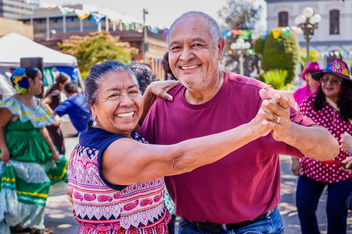 An older Latino couple smiling and dancing hand-in-hand at an outdoor festival.