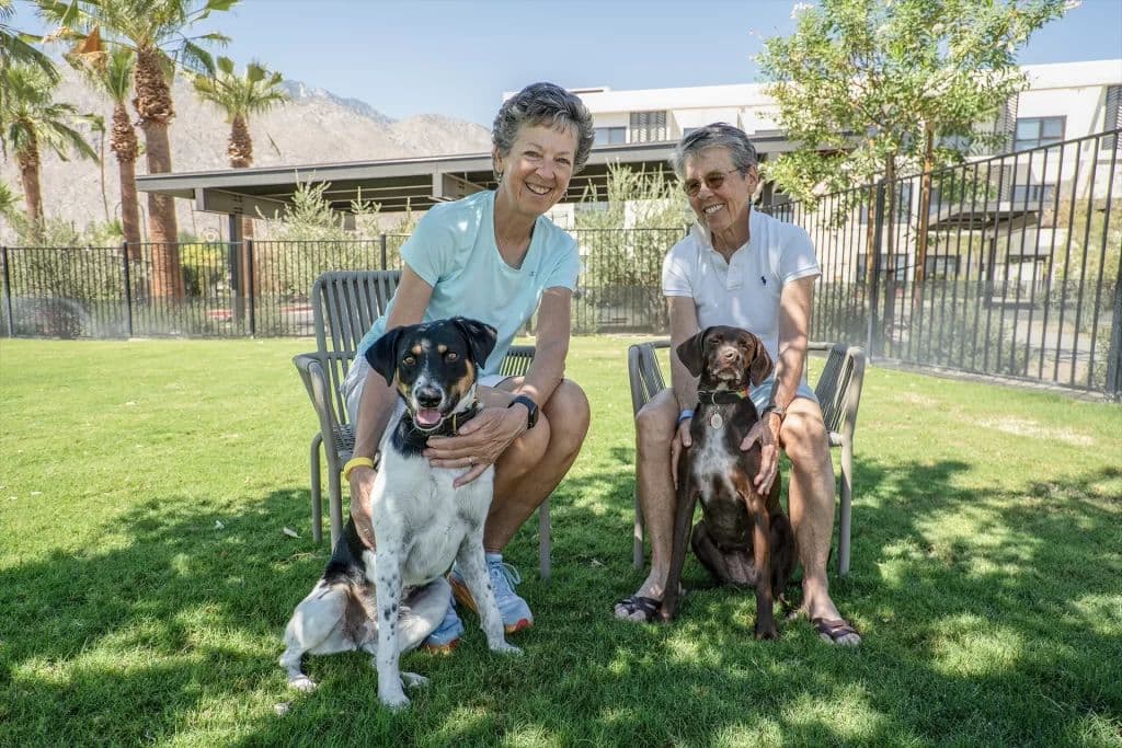 Two Living Out residents sitting on a lawn with their dogs, the community's buildings and the Palm Springs mountains behind them.