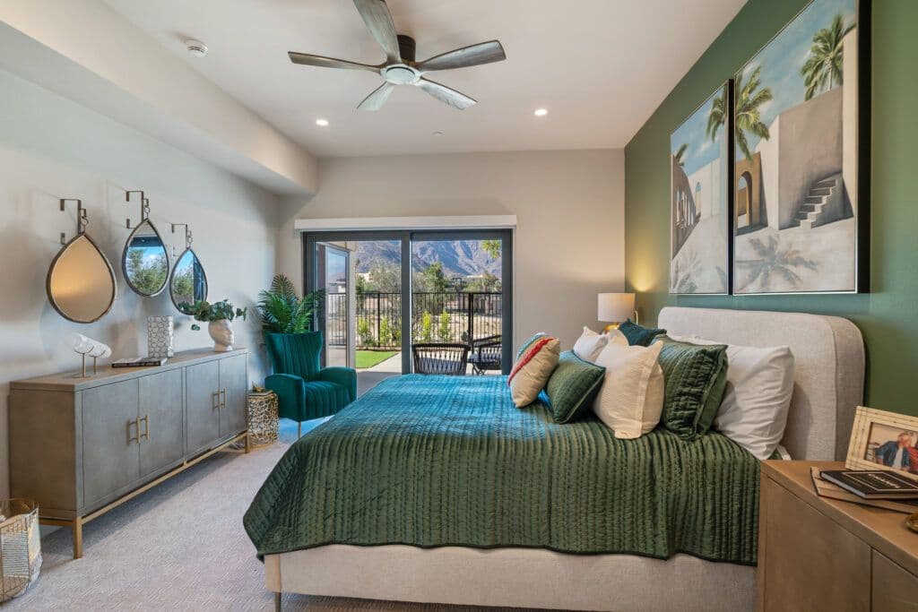 A bedroom in a Living Out residence, with a green quilted bed, a ceiling fan and sliding doors onto a patio with a mountain view.