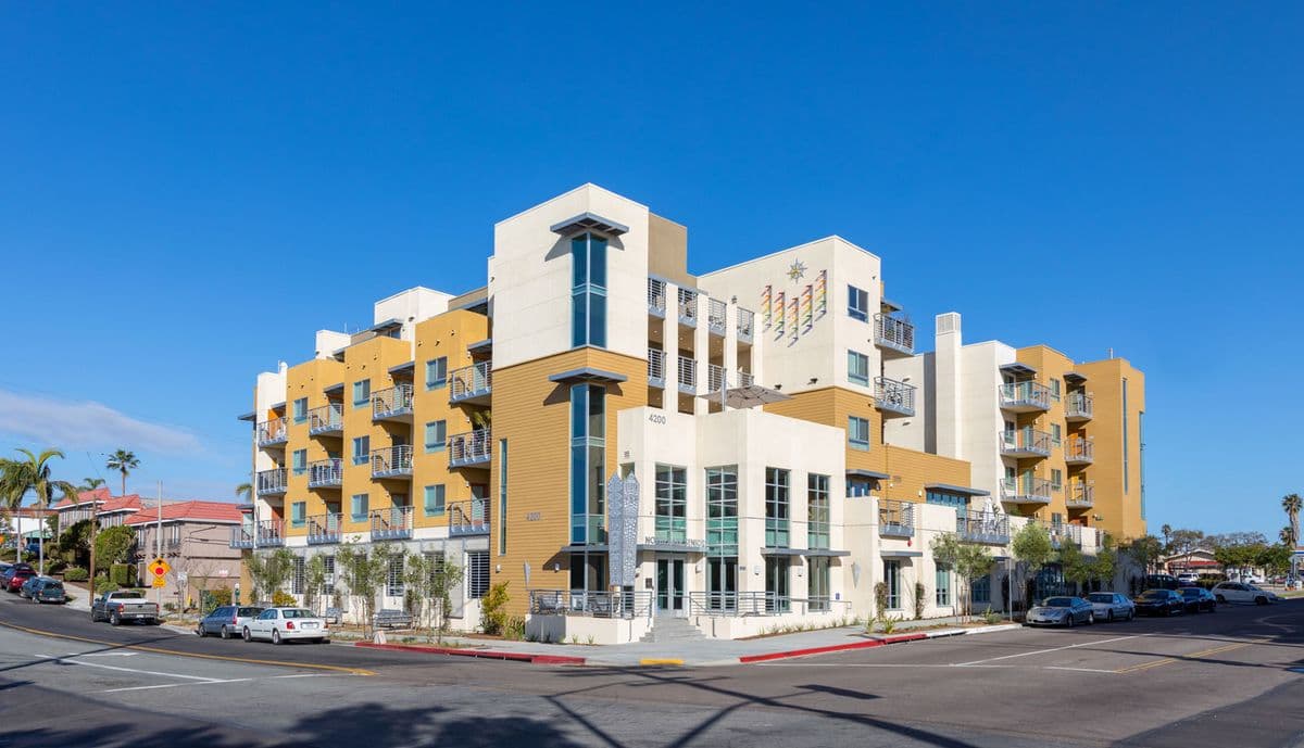 The four-storey North Park Seniors building on a corner in San Diego, cream and ochre with balconies and a rainbow sunburst artwork on the upper wall.