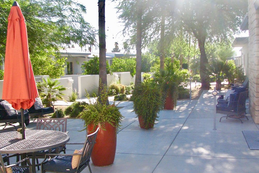 A shaded courtyard at Stonewall Gardens with patio dining tables, a folded orange umbrella and tall terracotta planters.